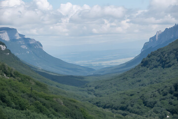 Obraz premium Expansive view of a deep, forested valley leading to a distant plain, framed by towering peaks