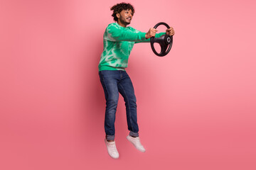 Young male model wearing green tie dye sweater jumps in mid air holding a steering wheel against...