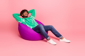 Young man relaxing on pink background sitting on a purple bean bag wearing green tie dye sweater jeans and white sneakers smiling
