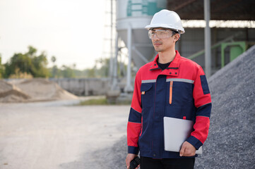 Asian male engineer in safety uniform holding a laptop and walkie-talkie at a concrete plant. Concept of site management, digital technology, and industrial work.