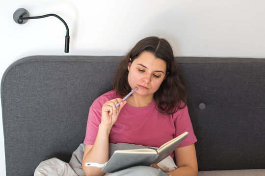 A woman sits in bed thoughtfully holding a pen and writing in her notebook, capturing a calm moment of journaling, daily planning, and personal reflection in a peaceful morning routine.