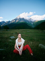 Fototapeta premium Meadow and mountains backdrop with expansive nature and clear sky, featuring a relaxed woman in casual wear sitting on grass, capturing a scenic outdoor moment of calm and style