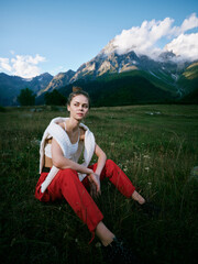 Fototapeta premium Woman sitting in a grassy meadow with towering mountains in the background, wearing a white top and red pants, calm mood, natural light and expansive alpine scenery for fashion and outdoor lifestyle