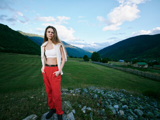 Fototapeta premium Woman outdoors on a grassy field with mountains in the background, wearing red pants and white top with a vest, casual fashion amid a scenic landscape and blue sky