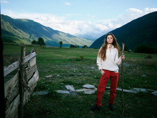 Fototapeta premium Girl standing with a wooden stick in a rural meadow, mountains in the distance and a rustic fence, nature scene capturing outdoor adventure and countryside beauty