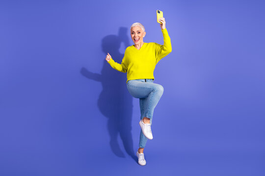 Elderly woman in bright yellow sweater takes joyful selfie while dancing against a vibrant purple background - Powered by Adobe