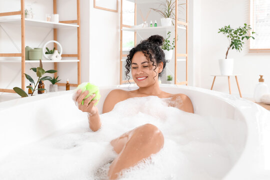 Beautiful young happy African-American woman taking bath with sponge and soap foam at home