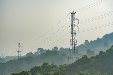 Electricity transmission towers and power lines stretching across a hazy, green forested mountain landscape
