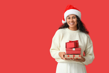 Happy African-American woman in Santa hat and with Christmas gifts on red background