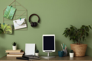 Stylish workplace with tablet on stand, books and different houseplants near green wall at home