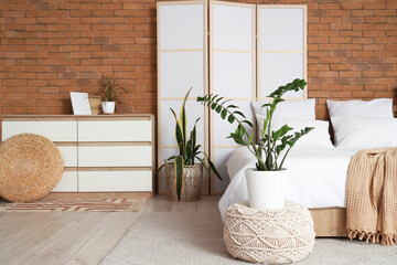 Interior of light bedroom with cozy bed, houseplants and white folding screen
