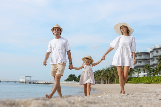 Happy family with daughter walking barefoot along beach during vacation, enjoying summer holiday time together, representing travel lifestyle, bonding and parenthood in tropical destination.