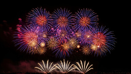 Colorful fireworks burst across the night sky above the city waterfront, reflecting on the water and creating a grand, vibrant celebration atmosphere.