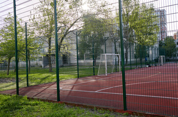 Сoated field for playing football behind the green fence mesh at school territory. Close-up of soccer field with red floor