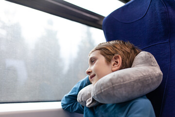 Young caucasian boy resting with travel neck pillow on express train, gazing out window at passing scenery. Comfortable travel.