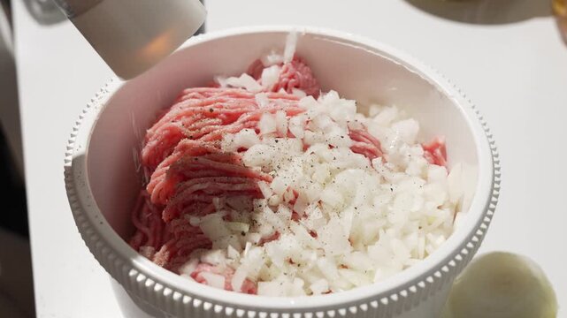 Salt is sprinkled over ground meat and diced onions in a mixing bowl. Classic seasoning for homemade patties, meatballs, and fillings.