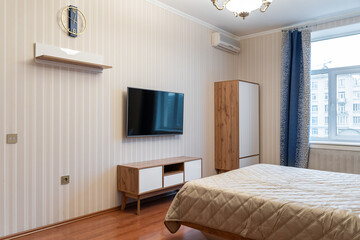 large, cozy bedroom with wooden bed, light-colored bedspread and pillows, blue curtains on window. bedroom has wooden chest of drawers, shelves and TV on wall. Retro chandelier on the ceiling.
