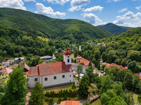 Aerial view of Church of St. Luke with its red roof nestled amid verdant trees and rolling hills, a peaceful scene under a bright sky., Sklene Teplice, Banska Bystrica Region, Slovakia.