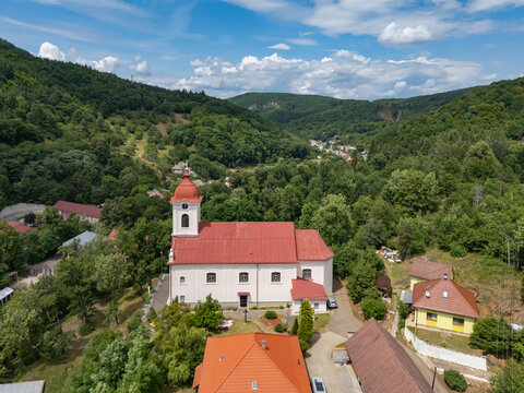 Aerial view of the Church of St. Luke with its vibrant red roof nestled amidst lush green trees under a cloudy sky, Sklene Teplice, Banska Bystrica Region, Slovakia.