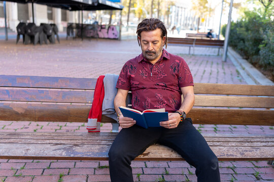 Middle-aged man focusing on a book while sitting on a wooden bench outdoors in the city