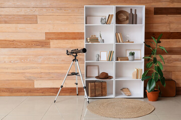 Bookshelf with decor, telescope and plants near wooden wall in room © Pixel-Shot