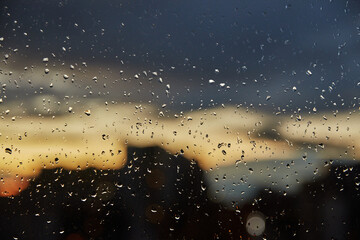 Moody view of an urban skyline silhouette through a rain-streaked window at dusk, featuring dramatic dark clouds and warm sunset light.