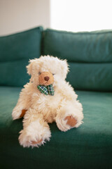 Soft plush teddy bear with a bow tie sitting on a green sofa in natural daylight. Cozy childhood atmosphere and home comfort concept. Vertical
