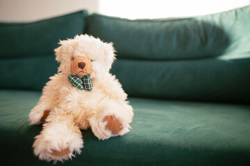 Soft plush teddy bear with a bow tie sitting on a green sofa in natural daylight. Cozy childhood atmosphere and home comfort concept.