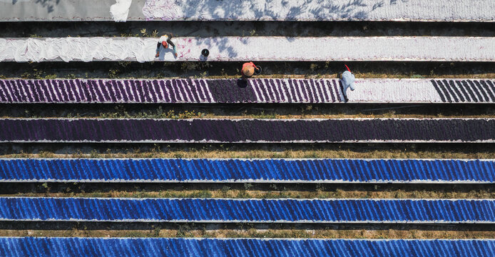 Aerial view of vibrant textiles laid out in neat rows, creating a colorful patchwork against the earth, as workers tend to the drying fabrics, Surakarta, Central Java, Indonesia.