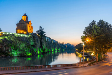 Night view of the old district of Tbilisi. The church and rocks are reflected in the Kura River and illuminated with colored lights. Blue sky.