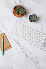 Top view of white keyboard, notebook and cactus on marble background in vertical minimalist layout with negative space. Concept of modern workspace and lifestyle