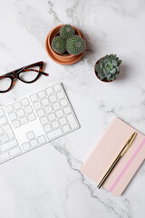 Top view of white keyboard, glasses, pink notebook and cactus on marble background in vertical composition with copy space. Concept of elegant workspace and creativity