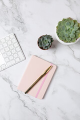 Vertical flat lay of succulents, white keyboard and pink notebook on a marble surface, minimalist and elegant workspace setup. Concept of modern workspace and creative lifestyle