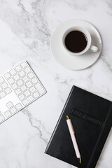 Top view vertical of coffee cup, white keyboard and black notebook on marble background, clean minimalist workspace style. Concept of modern workspace and creative lifestyle