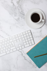 Top view vertical of coffee cup, white keyboard and turquoise notebook on marble background, minimalist and soft-toned workspace. Concept of modern workspace and creative lifestyle