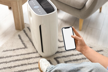 Young woman with mobile phone adjusting air purifier in living room, closeup