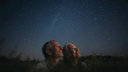 Gazing senior couple in casual clothes, tilting heads up, drawn by starry sky in tall grasses - Powered by Adobe