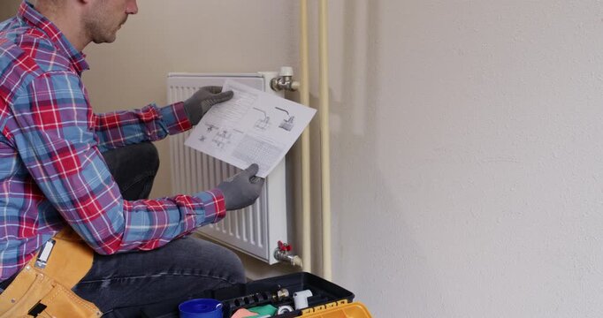 Male technician studies instruction sheet near radiator and toolbox on floor. Professional plumber compares diagram to fittings marking steps while torque values