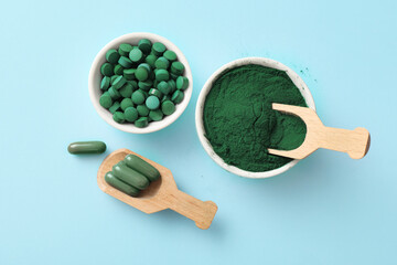 Bowls of spirulina tablets and powder with wooden scoops on blue background