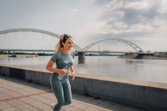 Young sporty woman running near river with bridge in background - Powered by Adobe