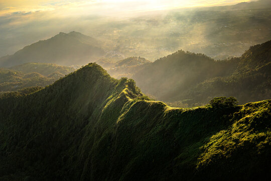 Aerial view of lush green mountains bathed in golden light, highlighting the ridge of Telomoyo Mountain under a soft, hazy sky, Magelang, Central Java, Indonesia.