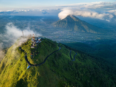 Aerial view of lush green mountains piercing through soft clouds, with winding roads leading to structures atop, Telomoyo Mountain, Magelang, Central Java, Indonesia.