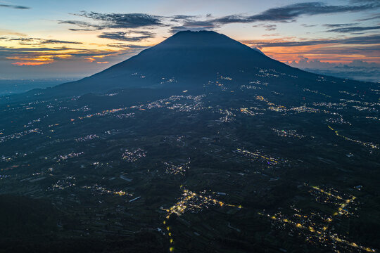 Aerial view of Merbabu Mountain under a dim sky, with scattered lights dotting the landscape below, creating a mesmerizing contrast of dark and light, Magelang, Central Java, Indonesia.