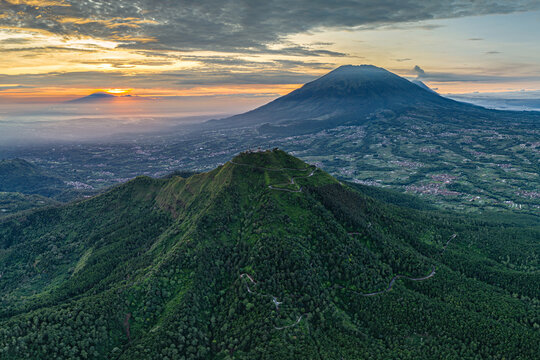 Aerial view of verdant Telomoyo Mountain under a warm sunset glow, with the majestic Mount Merbabu looming in the background, Magelang, Central Java, Indonesia.