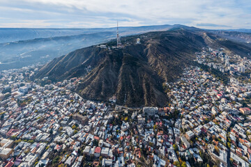 Aerial view of the city sprawling at the base of a prominent mountain capped with a radio tower, its slopes etched with paths of shadow and light, Tbilisi, Tbilisi, Georgia.
