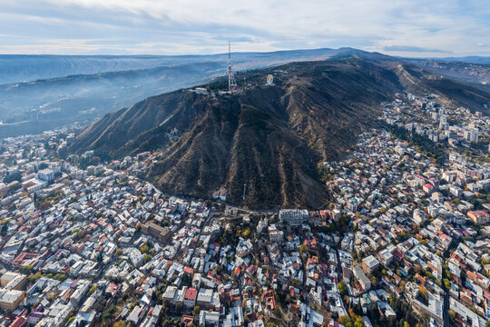 Aerial view of the city sprawling at the base of a prominent mountain capped with a radio tower, its slopes etched with paths of shadow and light, Tbilisi, Tbilisi, Georgia.