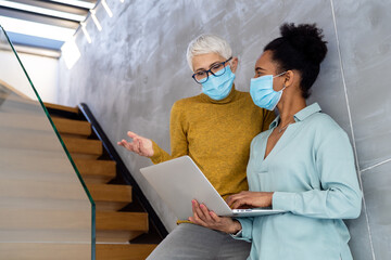 Women in startup workplace wearing masks, collaborating with laptop on creative projects in office