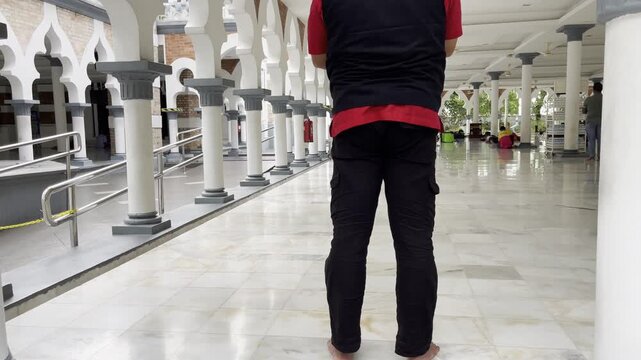 Anonymous man on knees stands up in prayer inside the serene marble corridor of Masjid Jamek Sultan Abdul Samad in Kuala Lumpur, surrounded by arches and white columns