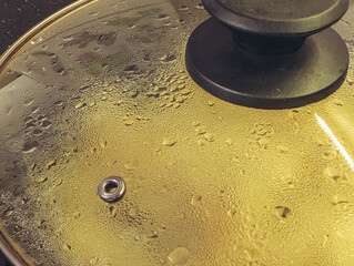Close-up of steam and droplets forming on a glass saucepan lid with a yellow light reflection.