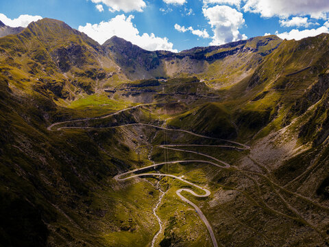 Aerial view of the winding Transfagarasan road snaking through the rugged, verdant mountains under a sky dotted with fluffy clouds, Curtea de Arges, Judetul Arges, Romania.